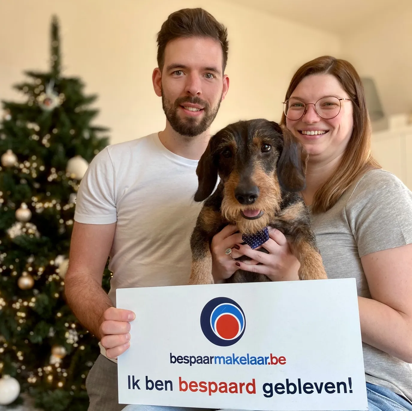 Koppel met hun teckel bij de kerstboom met het bordje 'Ik ben bespaard gebleven!'